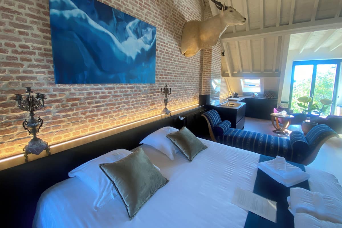 A hotel room with a brick wall, a mounted animal head, a large painting, a bed with pillows, a sofa, and a small table with a lamp.