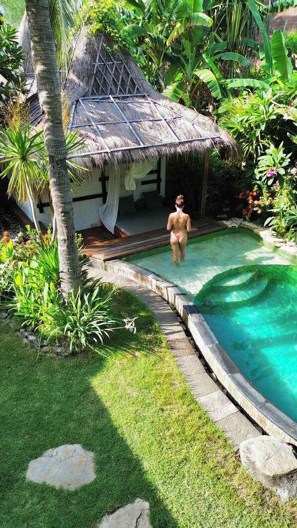 Eine Person geht auf einen Pool in einem tropischen Garten zu, mit einer strohgedeckten Hütte und üppiger Bepflanzung.