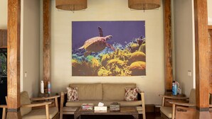 Un coin salon accueillant avec un banc en bois, des fauteuils confortables et un grand tableau représentant un paysage tropical.