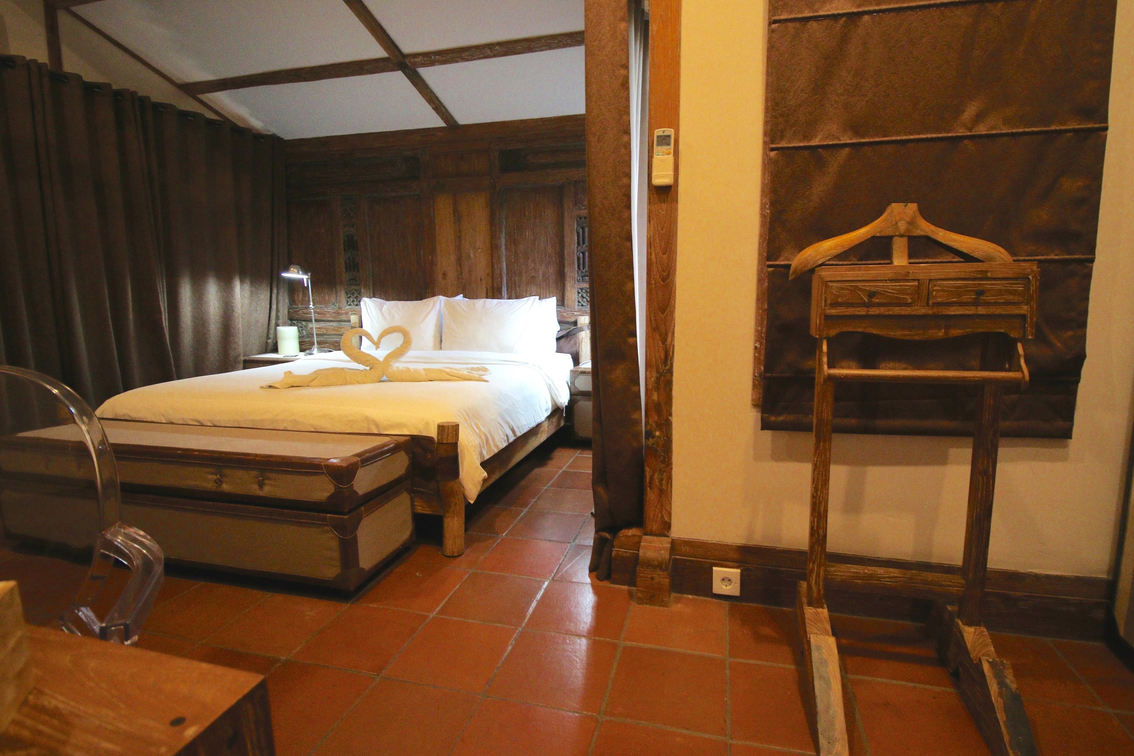 A hotel room with a bed, a wooden dresser, a suitcase, a wooden coat rack, and a lamp.