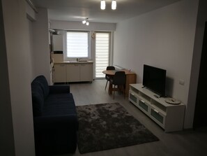 Ein Wohnzimmer mit einem dunkelblauen Sofa, einem Fernseher auf einem weißen Schrank, einem Esstisch mit Stühlen und einer Küchenzeile mit Fenster und Jalousien.