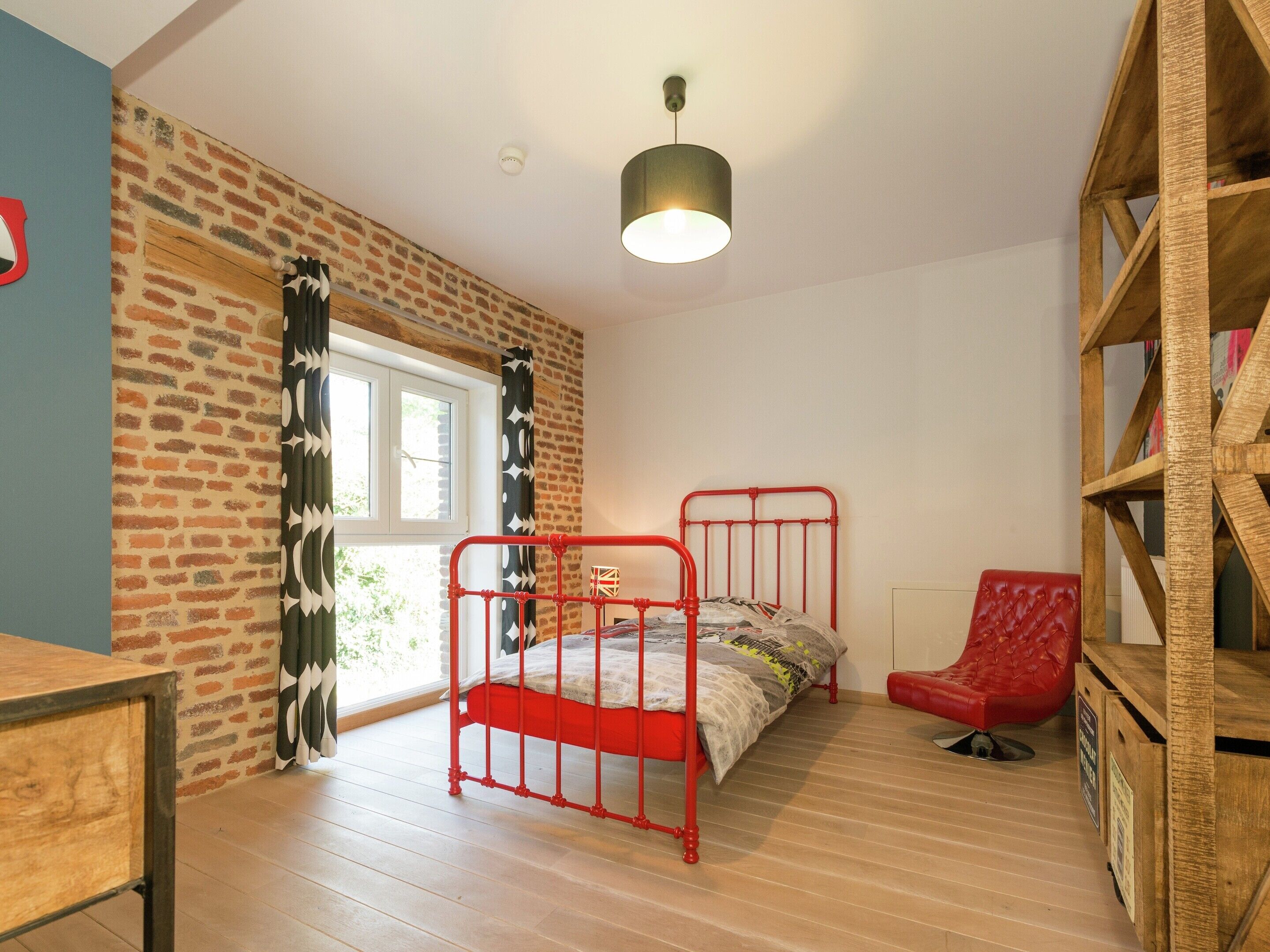 A room with a brick-patterned wall, a window with curtains, a red metal bed frame, a red chair, and a wooden bookshelf.
