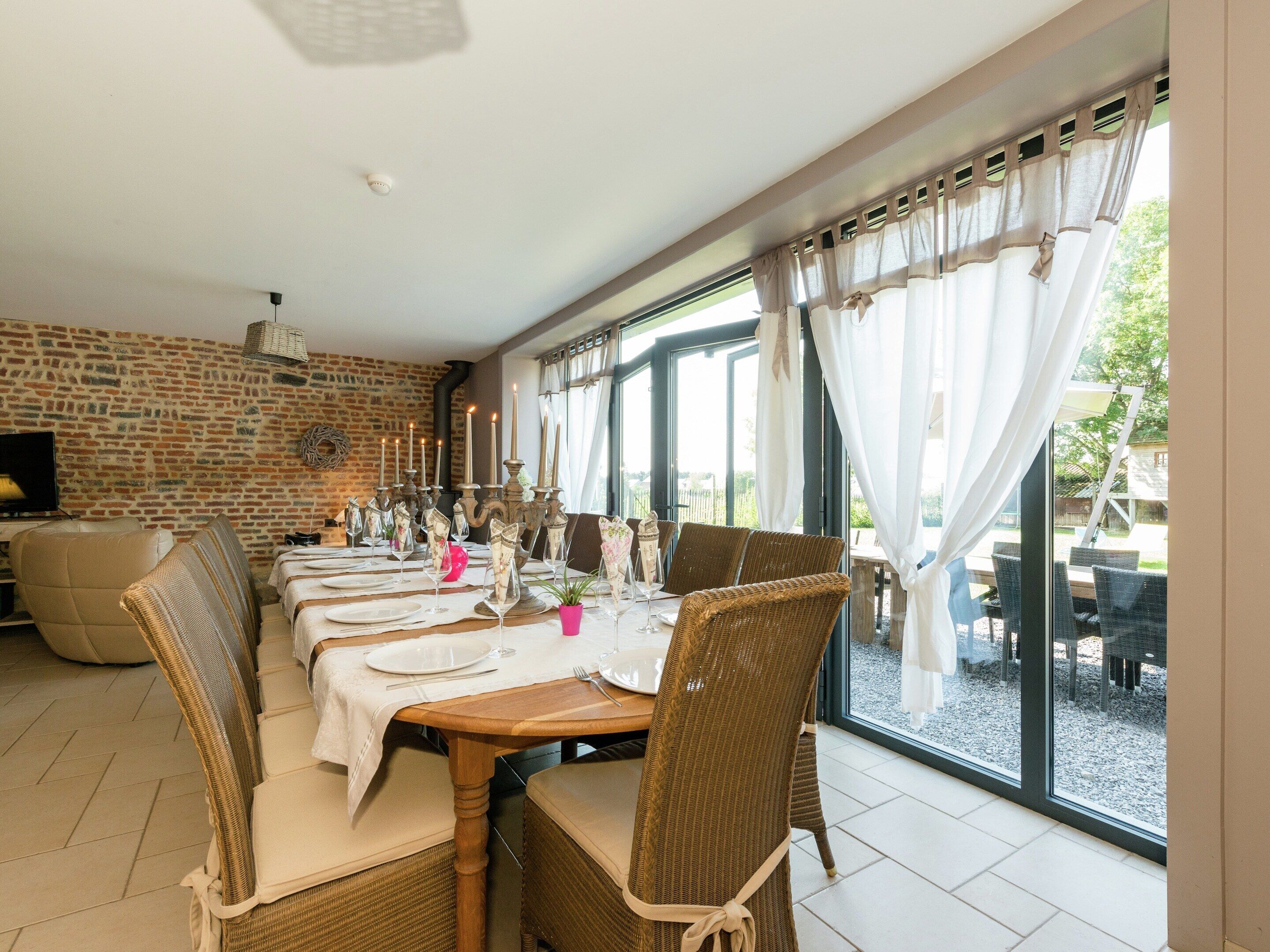 A dining area with a brick wall, wicker chairs, a wooden table set for a meal, and large windows with curtains.