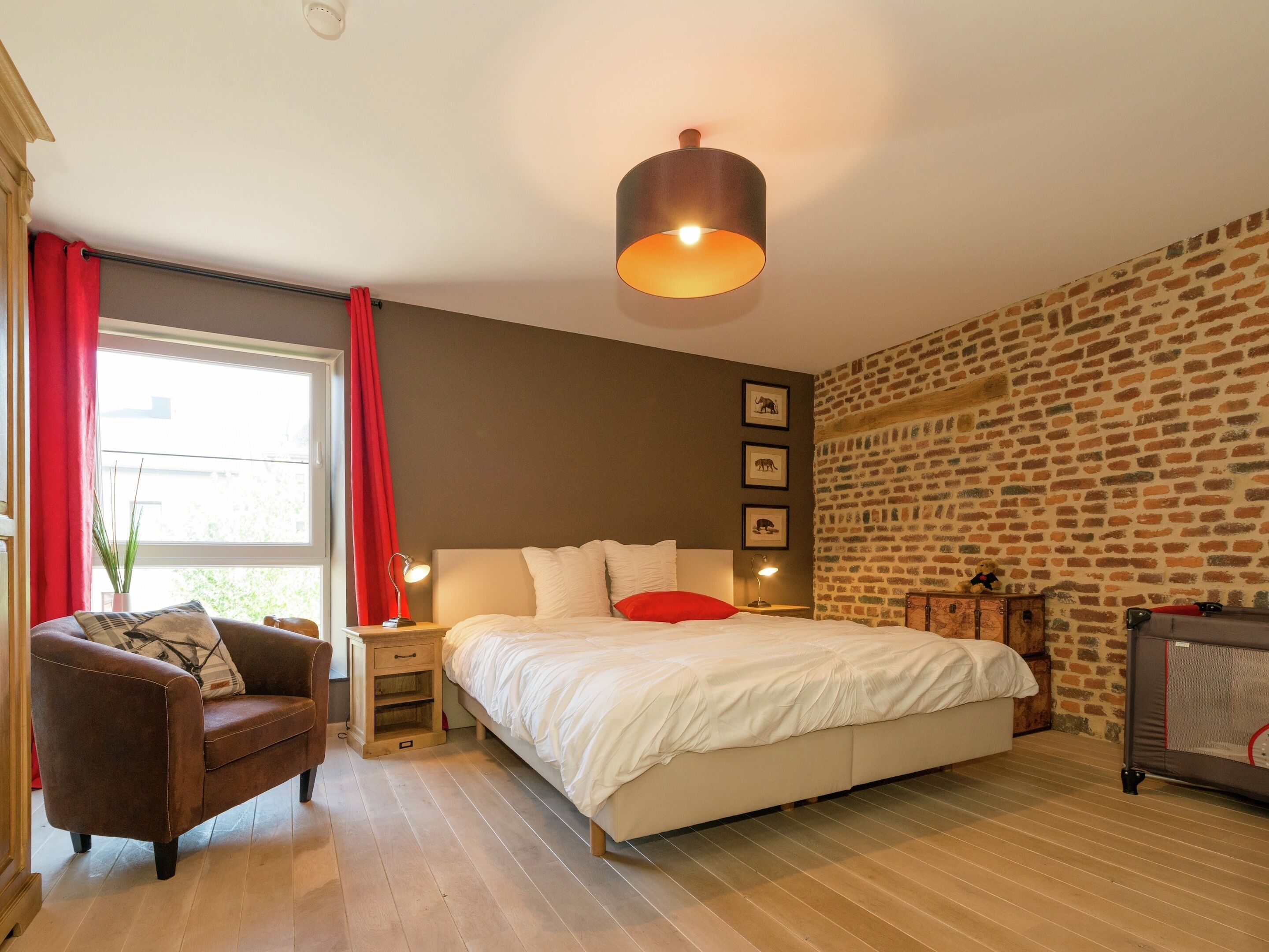 A bedroom with a large bed, a nightstand, a lamp, a chair, and a brick-patterned wall.