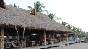 A beachside hut with thatched roofs, wooden furniture, and palm trees in the background.