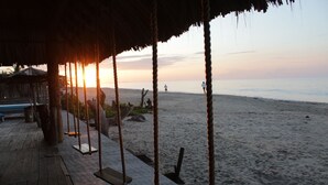 A wooden deck with swings and a thatched roof, overlooking a beach with a sunset.