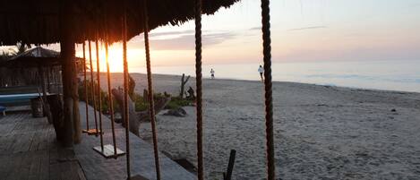 Una terraza de madera con columpios y techo de paja, con vistas a una playa al atardecer.