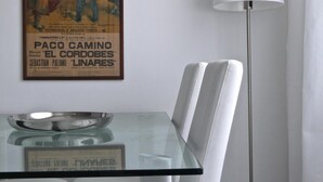 A modern dining area with a glass-top table, white upholstered chairs, a floor lamp, and a vintage movie poster on the wall.