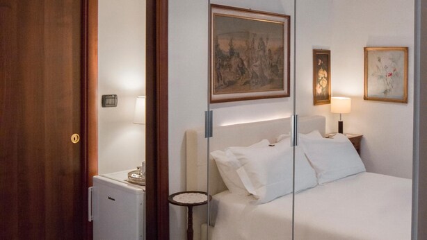 A hotel room with a bed, a small table, a lamp, a painting, and a door.