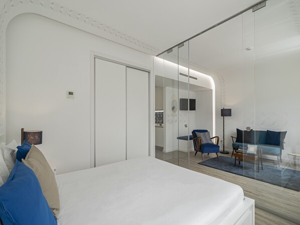 A modern bedroom with a large bed, a glass partition, a seating area with a sofa and chairs, and a small table.