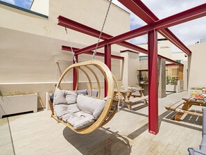 A hanging chair in a sunlit outdoor area with a wooden table and benches, surrounded by a red metal structure. - Drapes Design Apartments I (Funchal)