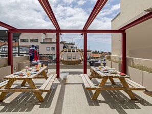 An outdoor dining area with wooden tables and benches, a hanging chair, and a view of the cityscape.