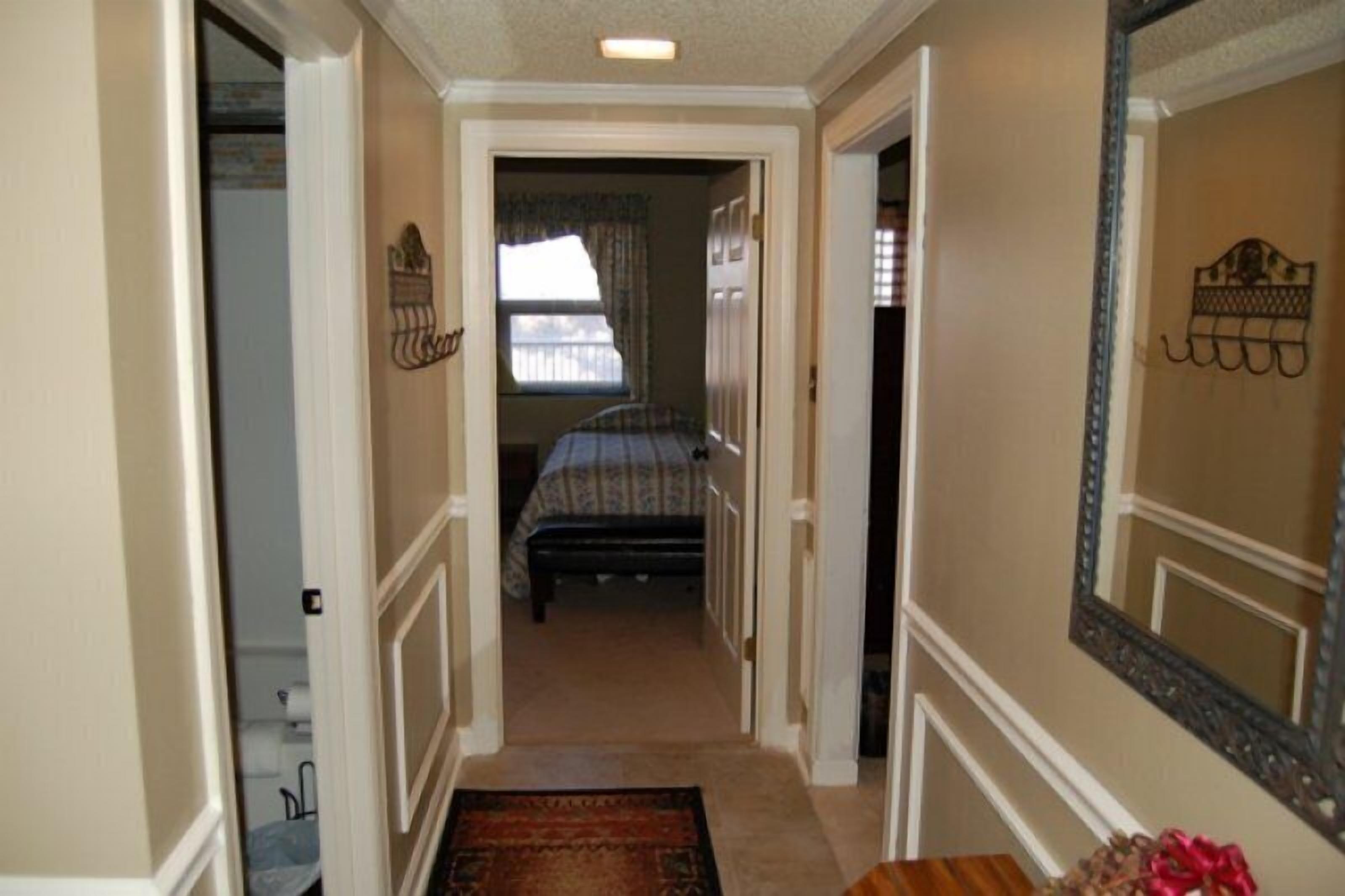 A narrow hallway leading to a bedroom with a bed, a window with curtains, and a large mirror on the wall.