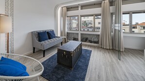 Un salon moderne avec un canapé, une petite table, un fauteuil en osier, une commode sombre et de grandes fenêtres avec des rideaux.