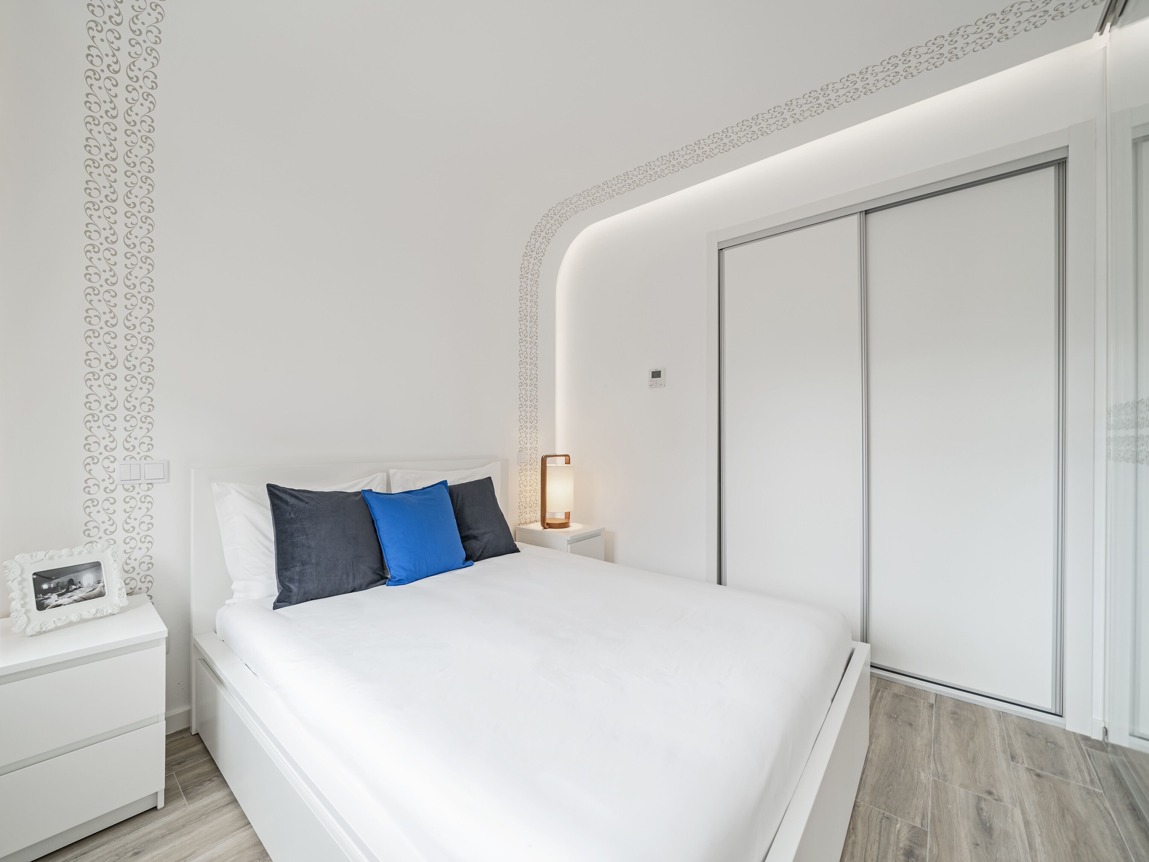 Un dormitorio moderno con una cama blanca, una mesita de noche, una lámpara y un armario.