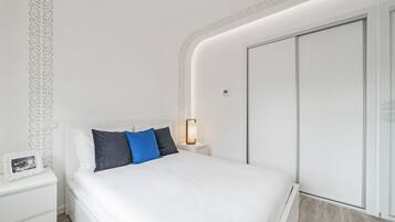 Un dormitorio moderno con una cama blanca, una mesita de noche, una lámpara y un armario.