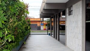 A covered walkway with a tiled floor, bordered by a low wall with dense green foliage. - Hotel Harmonia (Praia Grande)