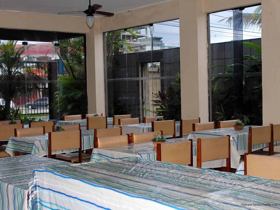 Un restaurante con varias mesas y sillas, amplias ventanas que dan a vistas de palmeras y edificios, y un ventilador de techo.