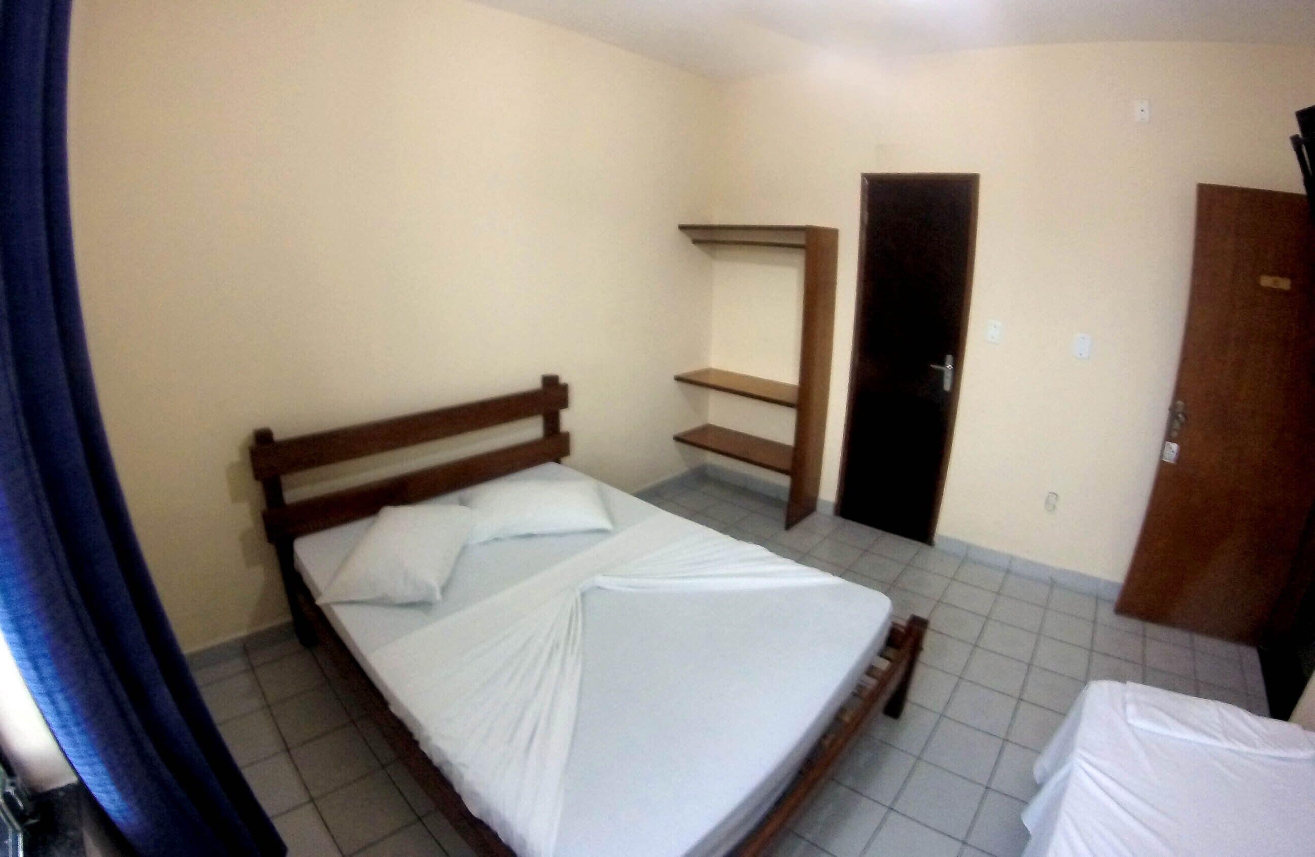 A small, simple room with a single bed, a wooden headboard, a small shelf, and a door.