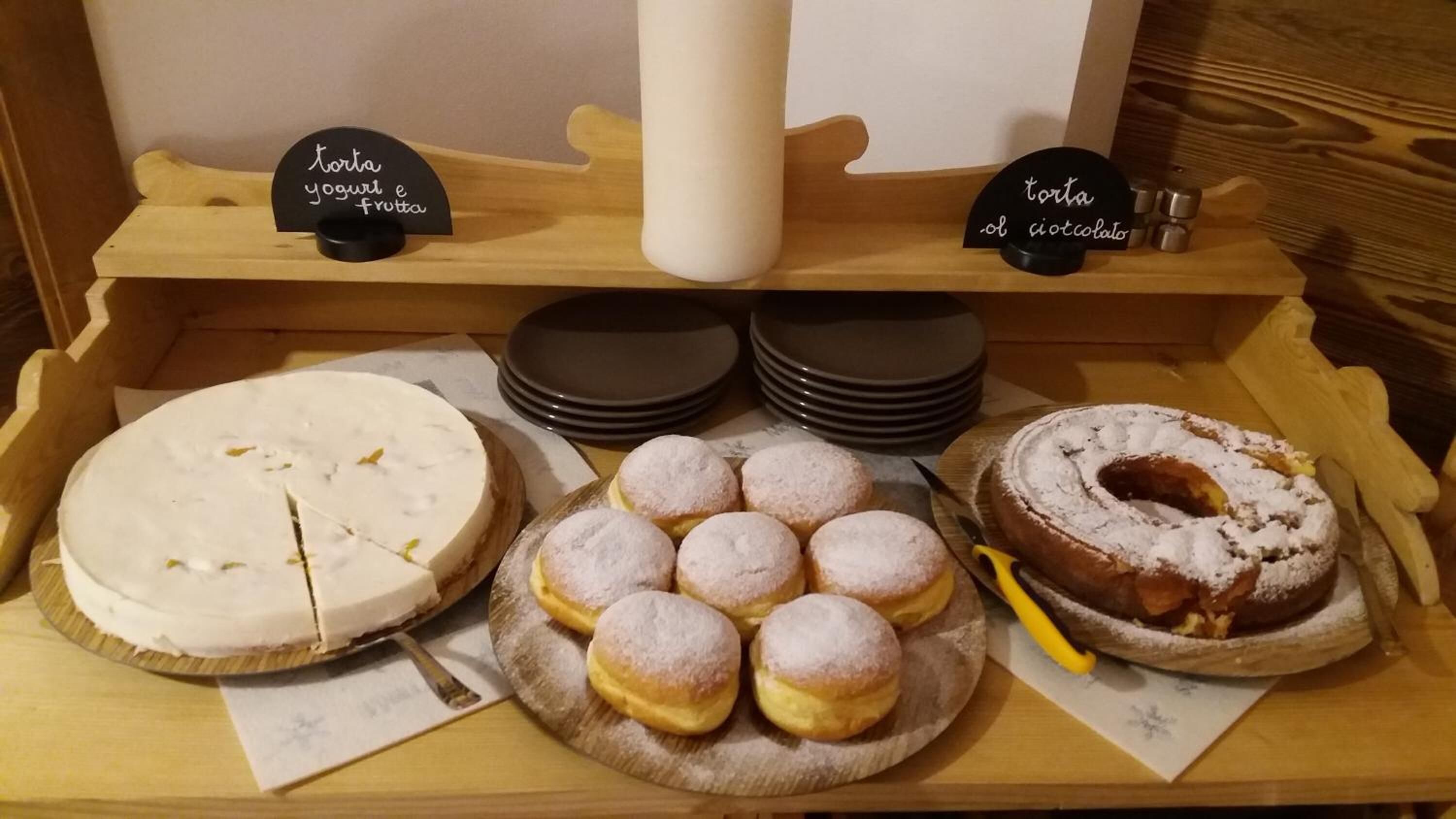 Esillä on erilaisia kakkuja ja leivonnaisia, kuten pyöreä kakku, josta on leikattu siivu, lautanen pieniä, pyöreitä leivonnaisia, jotka on peitetty tomusokerilla, ja rengasmaisen muotoinen kakku, jonka päällä on tomusokeria.