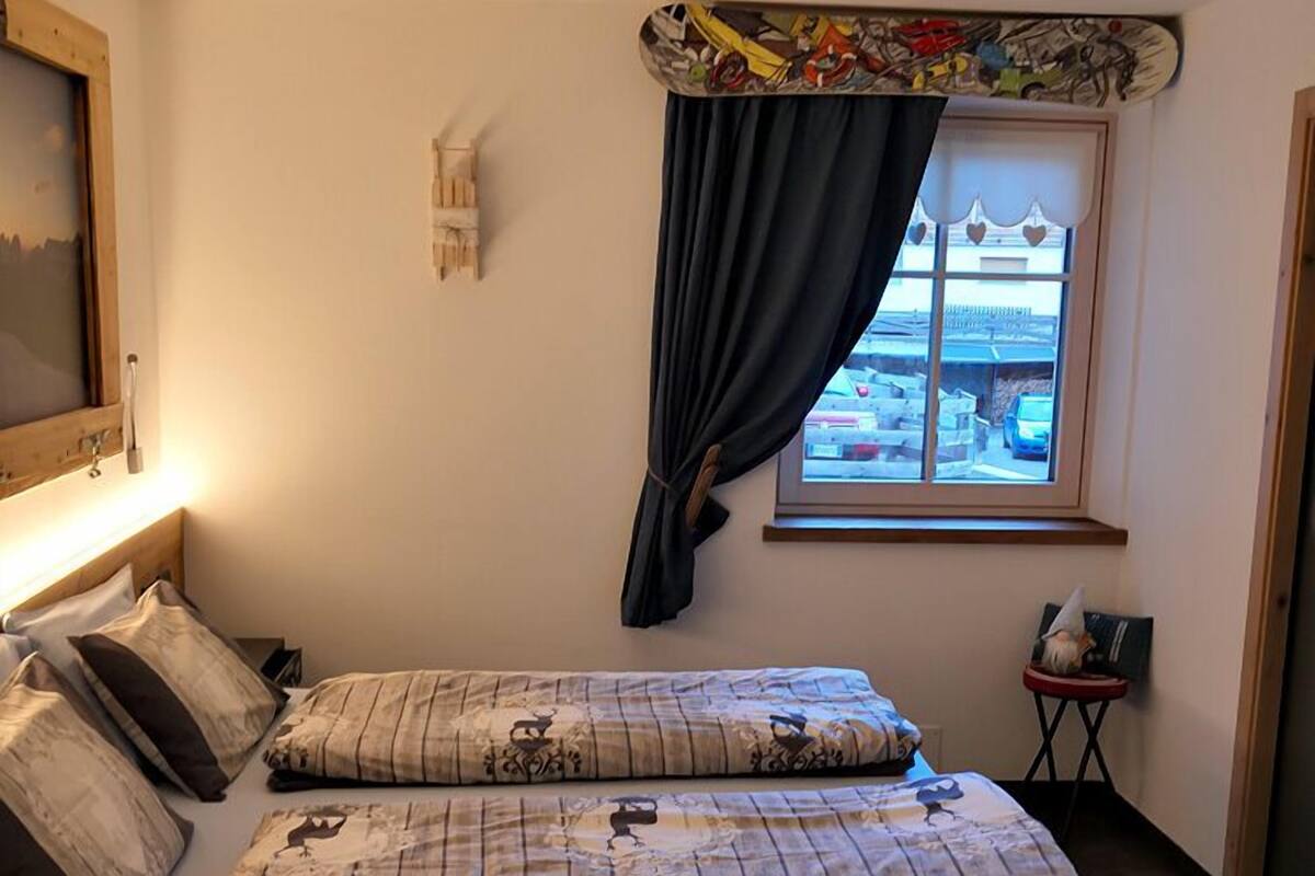 A bedroom with a bed, pillows, a nightstand, a window with curtains, and a wall-mounted light fixture.