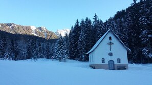 Una iglesia pequeña y blanca con una cruz en la fachada, rodeada de árboles cubiertos de nieve y montañas al fondo.