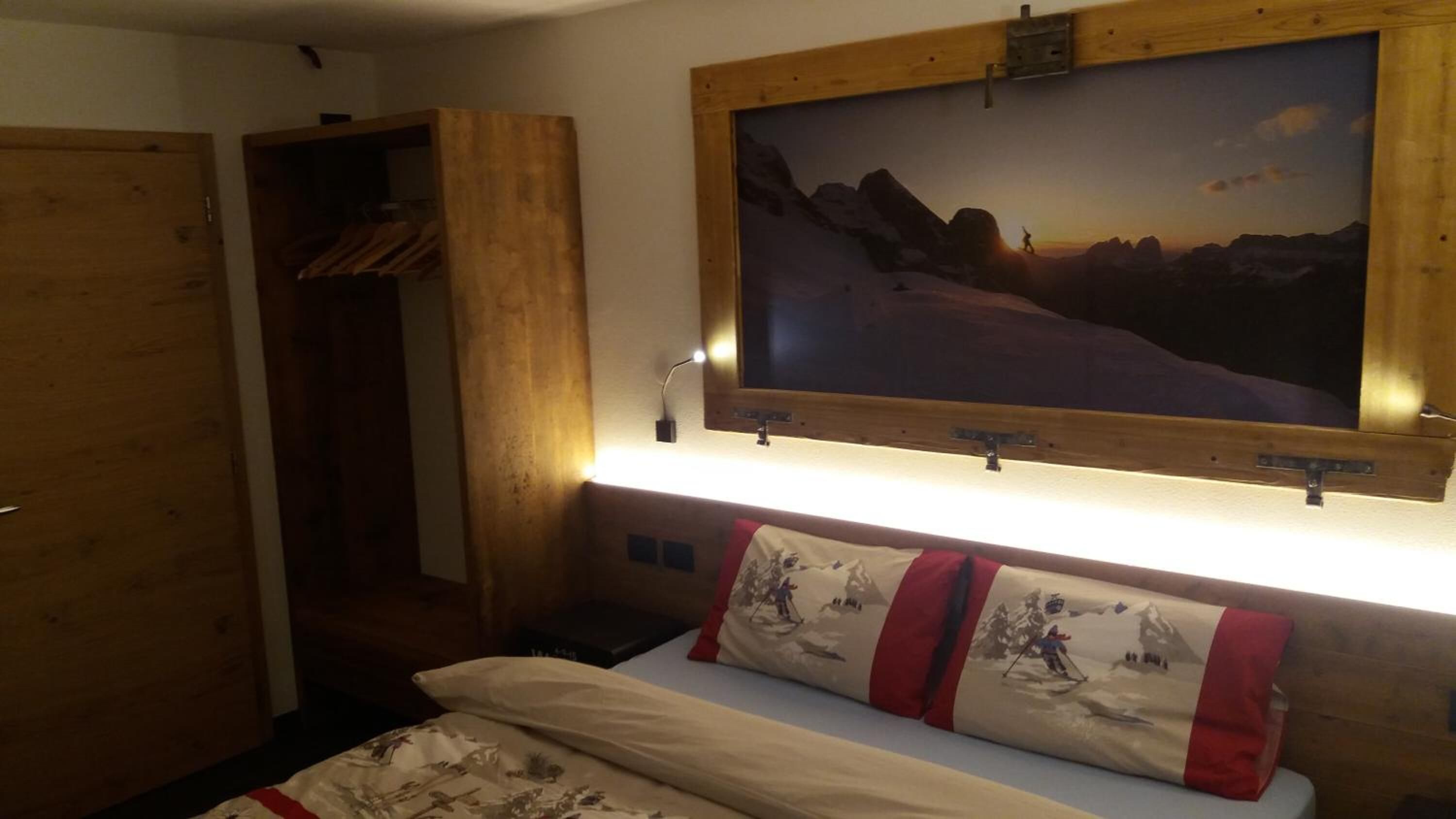 A bedroom with a bed, a wooden wardrobe, a mounted TV, and a painting of a mountain landscape.