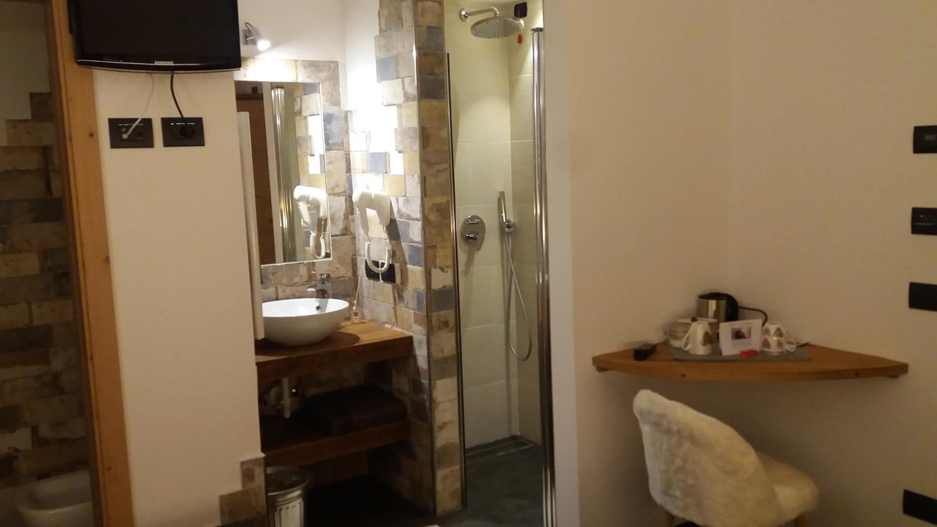 Un baño con pared de piedra, un lavamanos blanco, cabina de ducha con puerta de vidrio y una toalla colgada de un gancho.