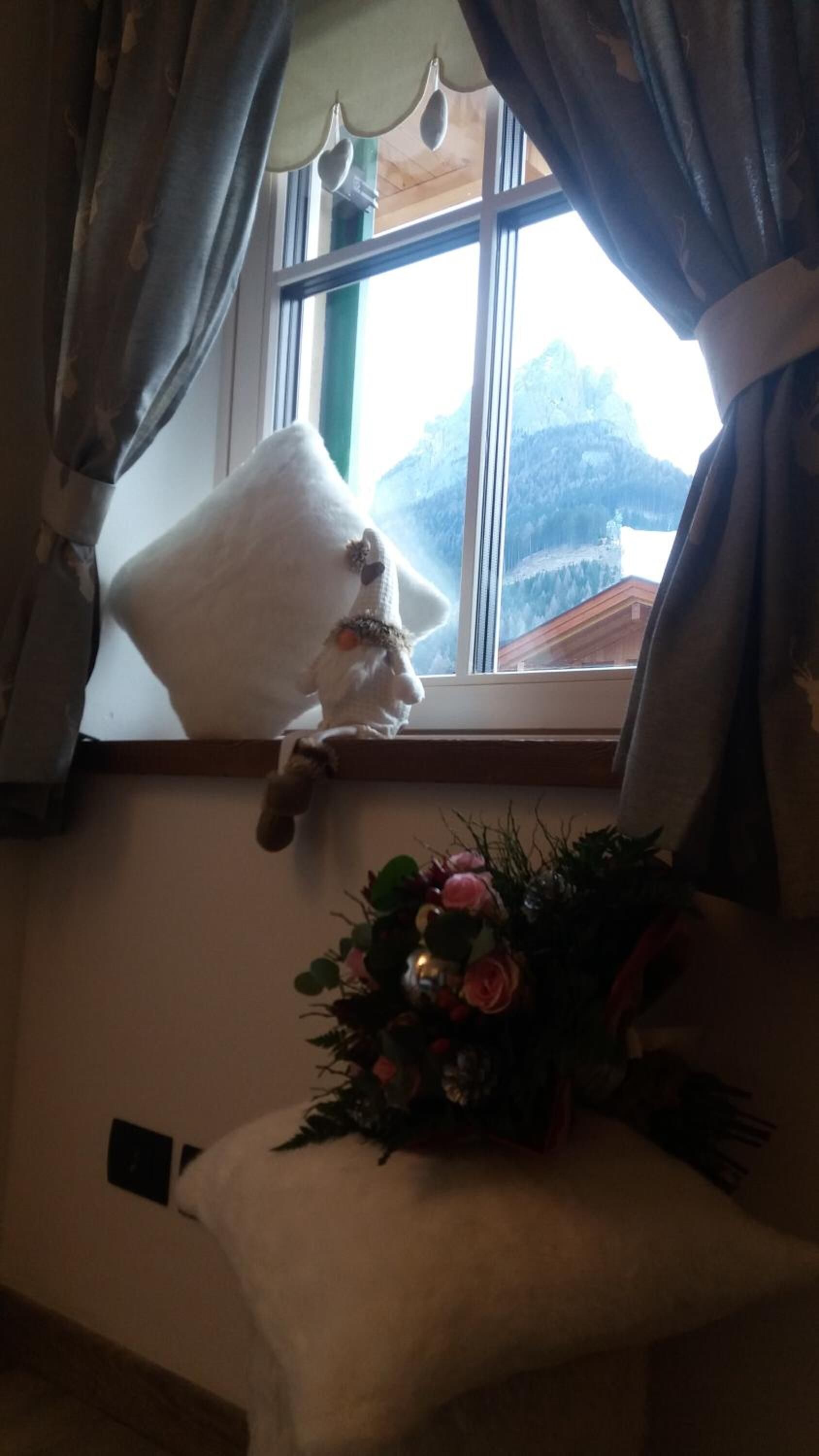 A room with a window, a plush white pillow, and a floral arrangement on a bedside table.