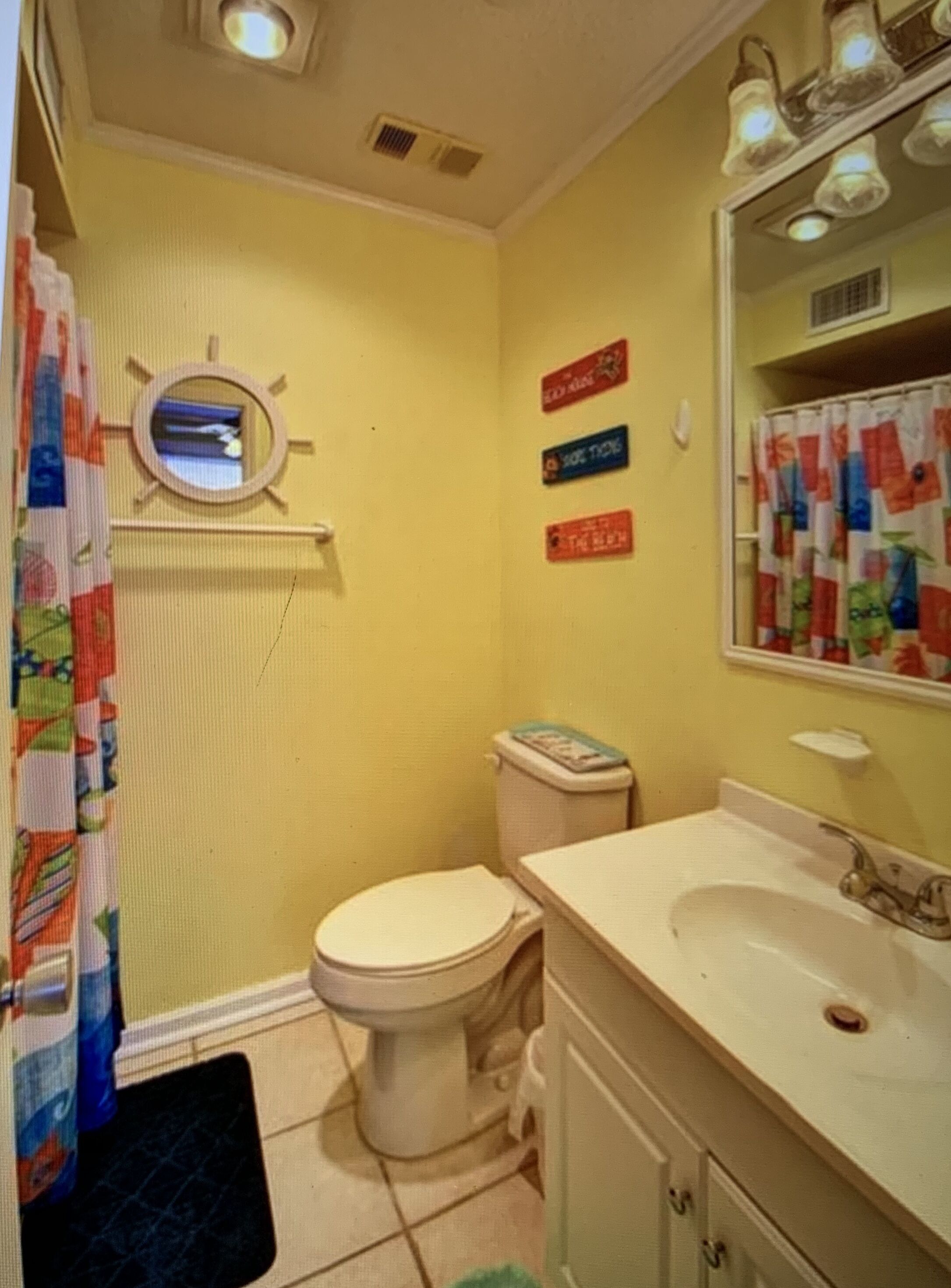 Une salle de bain avec des toilettes, un lavabo et un miroir. Des panneaux colorés sont accrochés au mur, et il y a un rideau de douche à motifs.