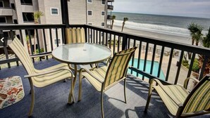 A balcony with a glass table and striped chairs, overlooking a beach and a swimming pool. - Beachfront/Pool Murrells/GardenCity/Surfside 
Wkly/MilitaryFirePoliceNurse Discs (Murrells Inlet)