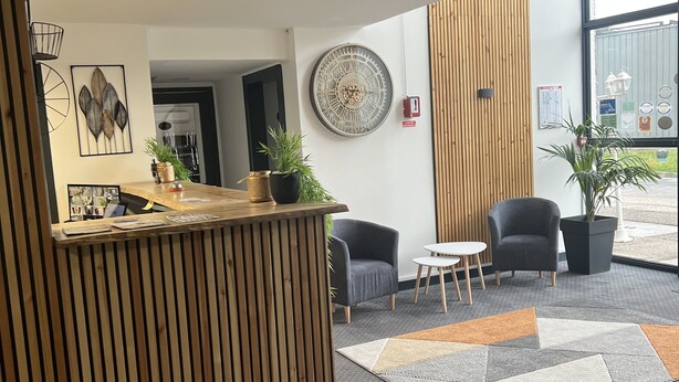 A reception area with a wooden reception desk, a large wall clock, and seating area with chairs and a small table. - Hotel de la Paix Lyon Sud Givors (Givors)