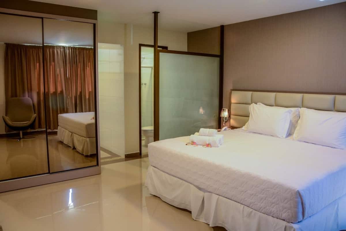 Une chambre d’hôtel avec un grand lit, une chaise, une cloison en verre et une salle de bain visible à travers cette cloison.