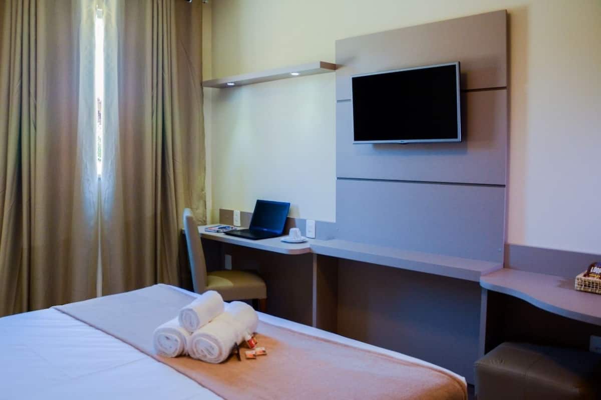 Une chambre d’hôtel avec un lit, un bureau avec un ordinateur portable, un téléviseur fixé au mur et une fenêtre avec des rideaux.