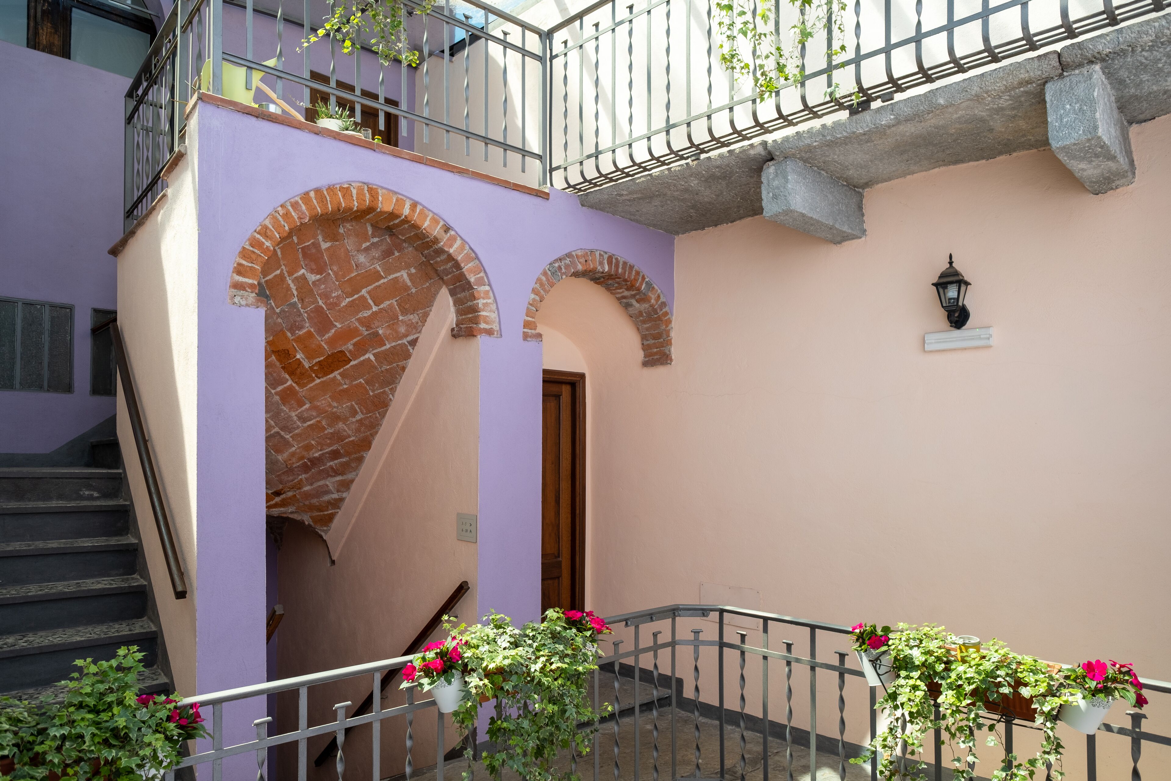 A building with a purple facade, brick arches, a staircase, and a balcony with potted plants.