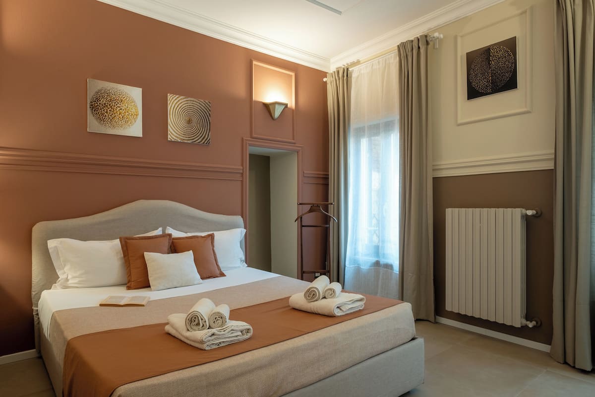 Habitación de hotel con una cama grande, una mesita de noche, un radiador y una ventana con cortinas.