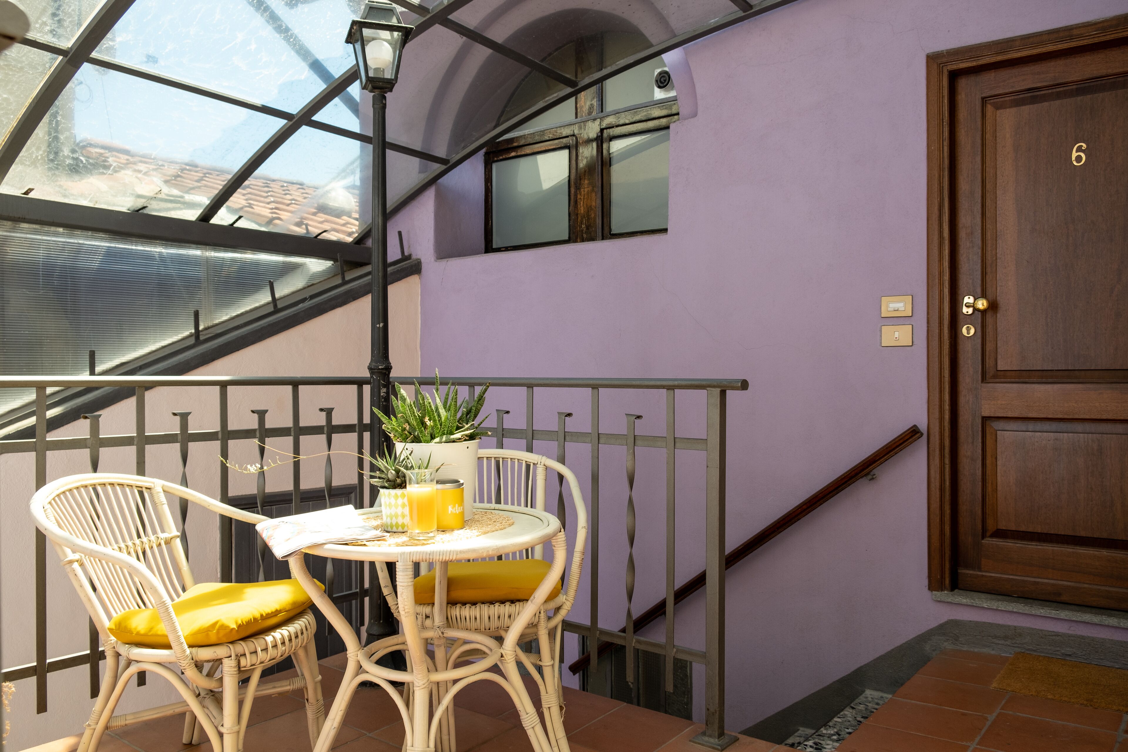 A balcony with wicker chairs and a small table, a wooden door with the number 6, and a view through a glass window.