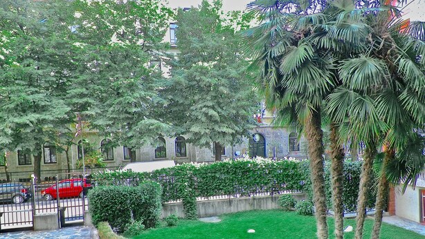 A courtyard with a stone pathway, green lawn, palm trees, and a red car parked near a gate. - Aiello Suites Executive (Milan)