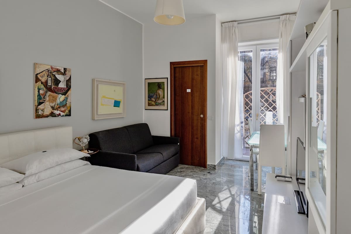 A modern hotel room with a bed, a sofa, a desk with a computer, and artwork on the walls.
