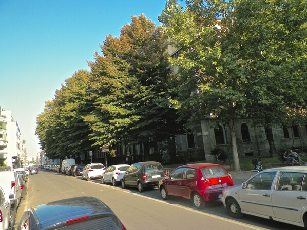Une rue bordée de voitures garées et d’arbres, avec un bâtiment aux fenêtres en arc de cercle en arrière-plan.