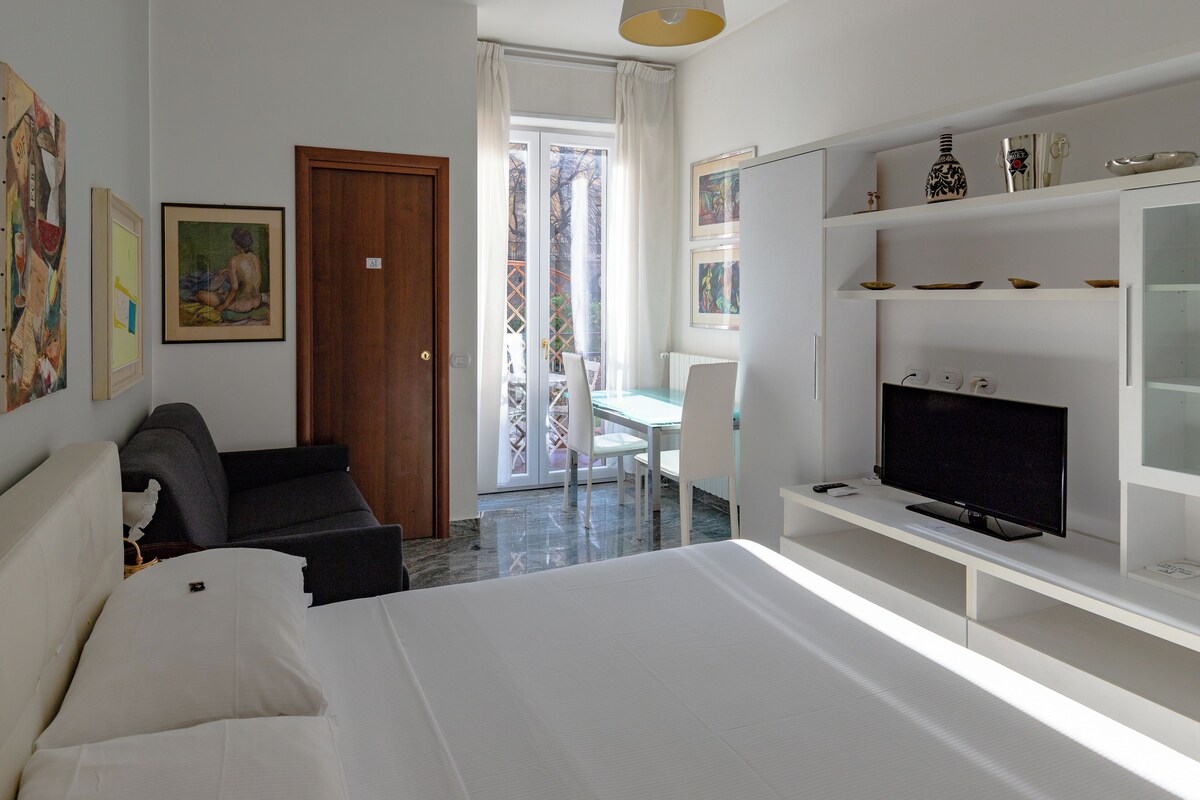 A hotel room with a bed, a TV, a desk with a chair, and artwork on the walls.