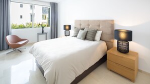 Een moderne slaapkamer met een groot bed, twee nachtkastjes met lampen, een stoel en uitzicht op groen buiten.