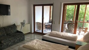 A modern living room with a sofa, a bed, and a television. There is a sliding glass door leading to an outdoor area with a table and chairs. - Gästehaus Denninger (Karlsbad)