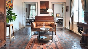 A room with a brown leather sofa, a round coffee table, a patterned rug, a wooden side table, and a large window with curtains. - Les Tuileries du Buisson (Verneuil d'Avre et d'Iton)