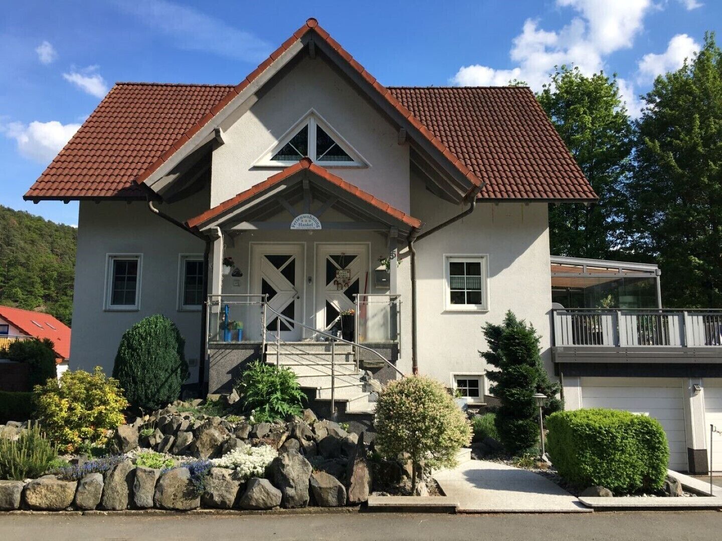 Uma casa de dois andares com telhado de telhas vermelhas, varanda frontal e um muro de suporte de pedra.