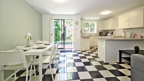 A modern kitchen with white cabinets, a black and white checkered floor, and a dining area with white chairs and a table set for two. - Harlinger Lodge Annexe (London)