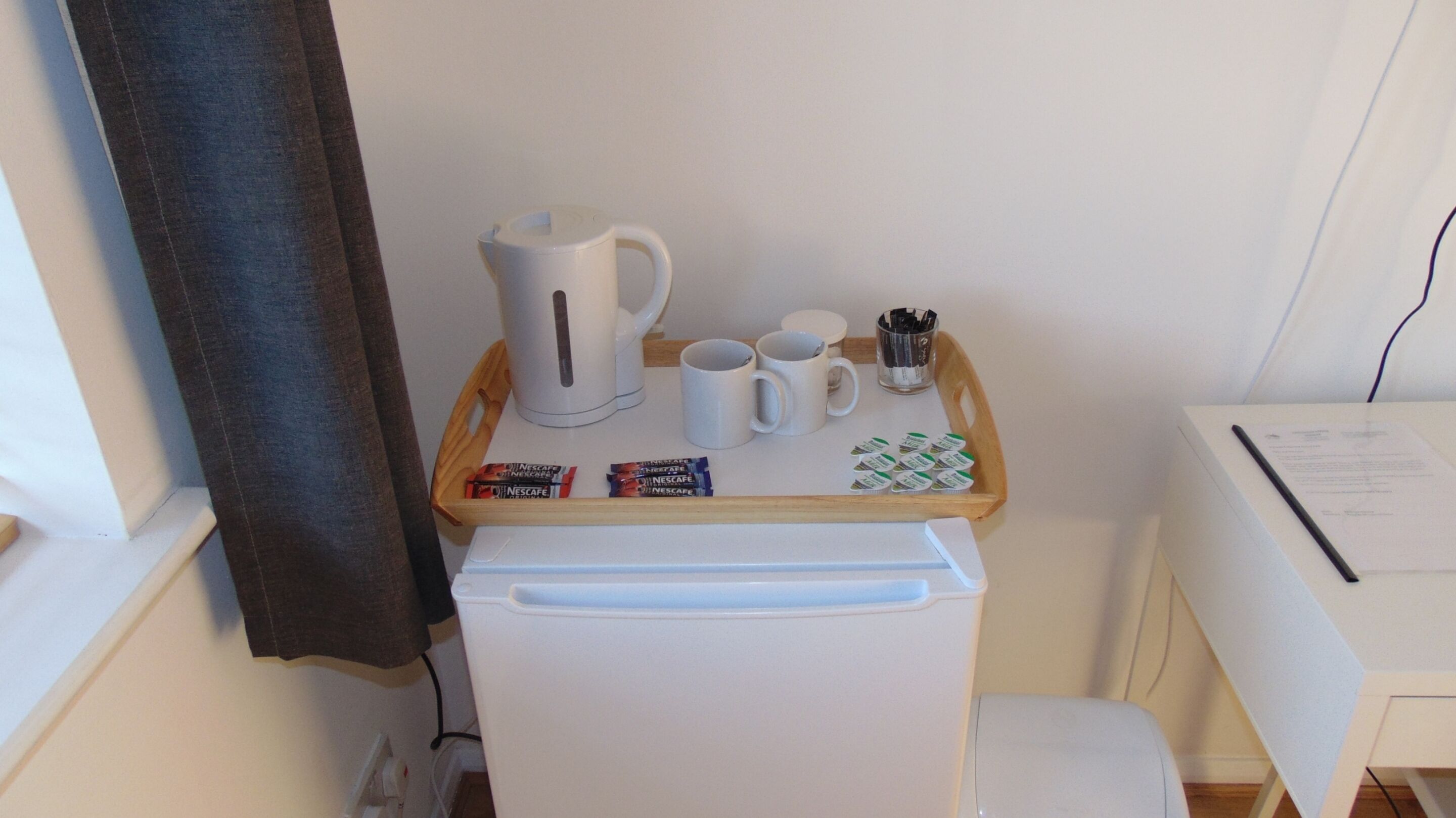 A compact kitchenette with a coffee maker, two mugs, a tray with coffee pods, and a small trash bin.
