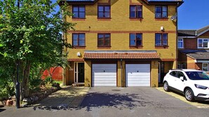A two-story building with a red-tiled roof, two garage doors, and a driveway with a parked white car. - Harlinger Lodge Annexe (London)