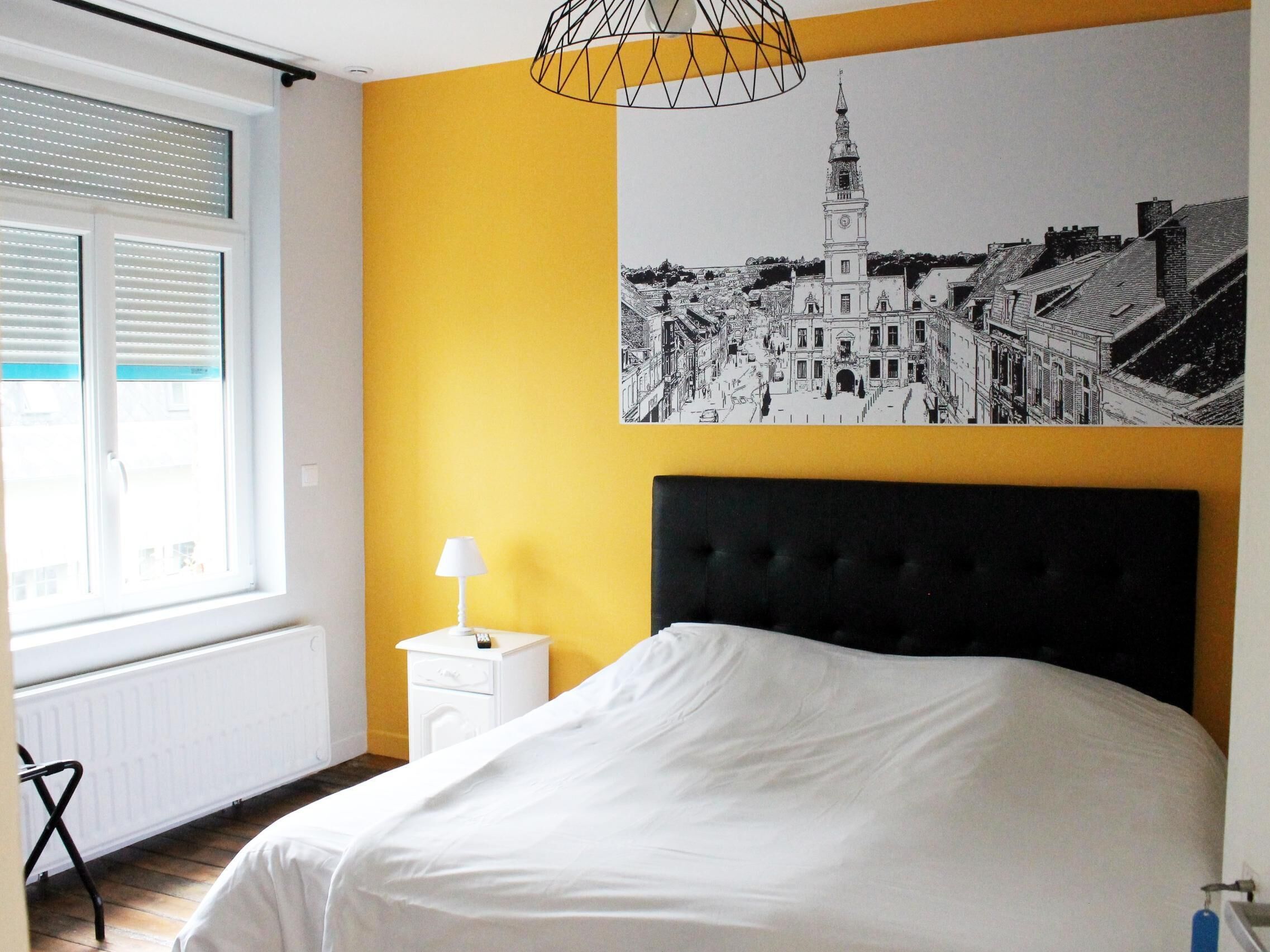 A bedroom with a bed, a nightstand, a lamp, a window with blinds, and a cityscape painting on the wall.
