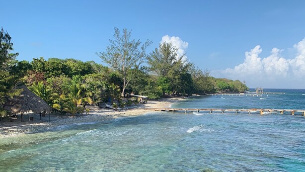 Ein Strand mit klarem türkisblauem Wasser, einem Sandstrand und einer kleinen Insel mit Vegetation und einem Gebäude mit Strohdach.